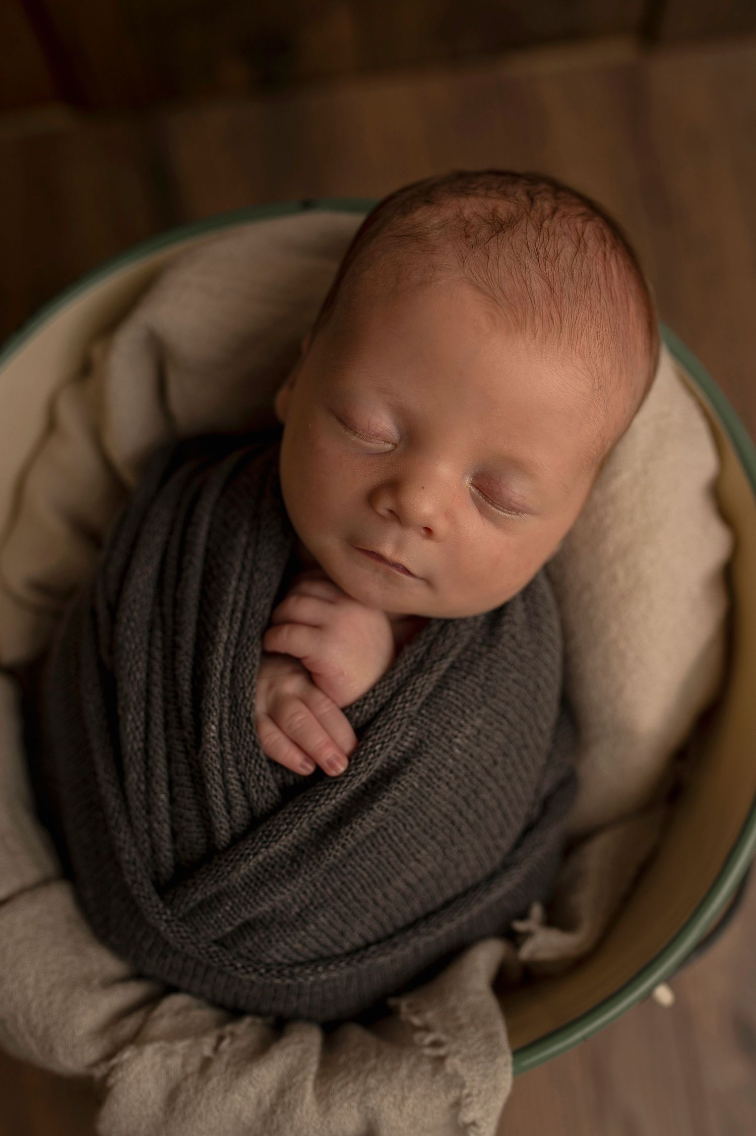 Close-up of peaceful newborn baby swaddled in gray knit wrap resting in round cushioned container.