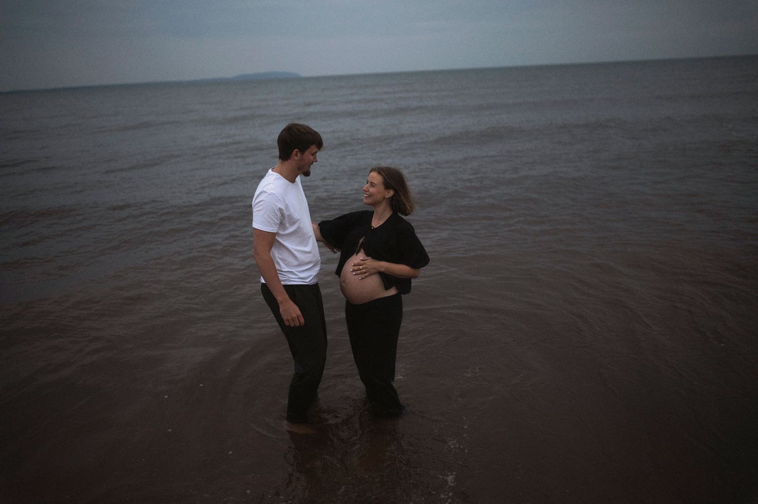 Couple standing in shallow water during maternity photoshoot at dusk.