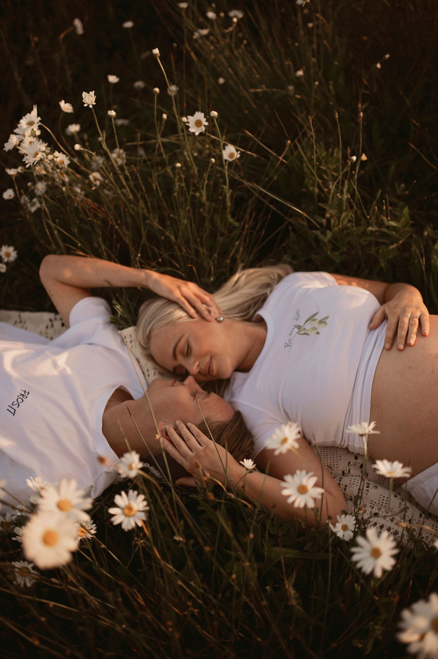 A couple wearing white clothing lies together in a field of blooming daisies at dusk.