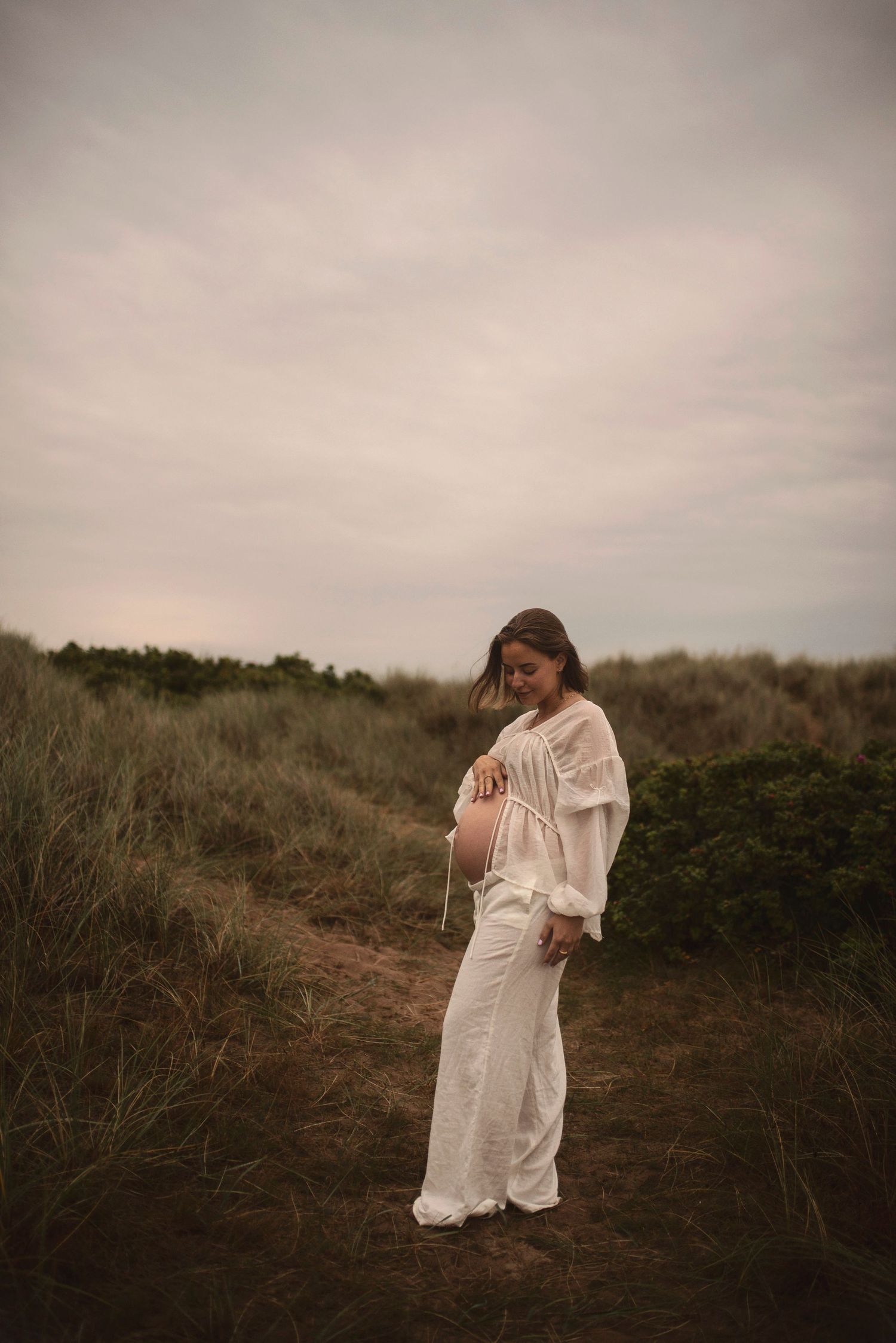 Expectant person in white outfit stands on natural path among coastal grasses.