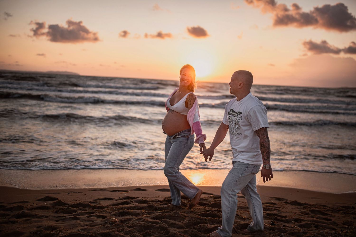 A couple walking hand in hand on a sandy beach during golden hour sunset as waves roll in from the ocean.