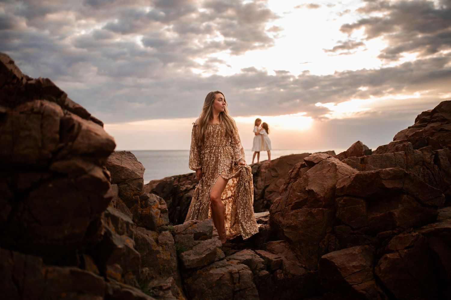 Person in flowing dress stands on rocky coastline during dramatic sunset.