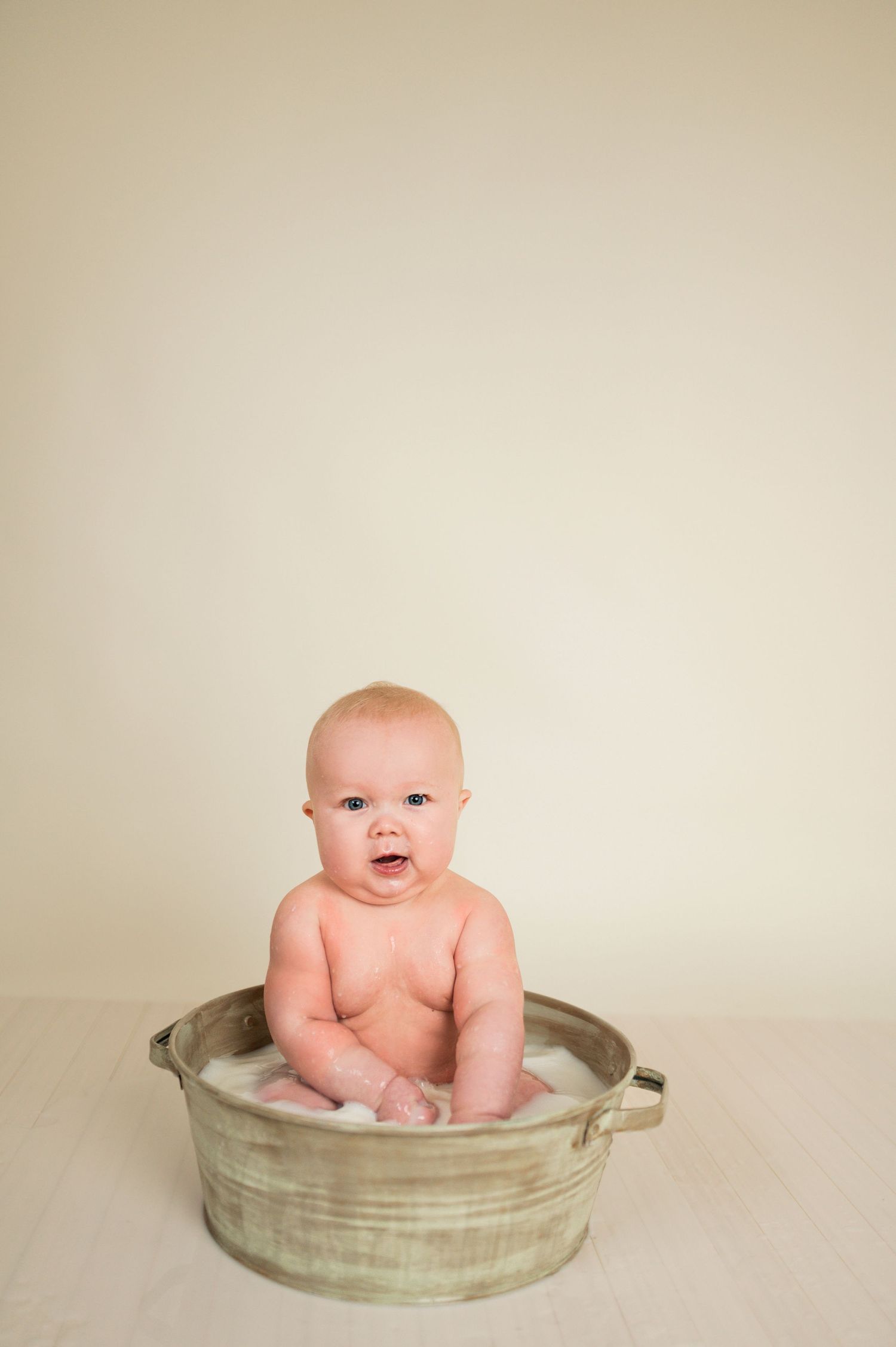 Baby sitting in vintage metal wash basin against neutral backdrop.