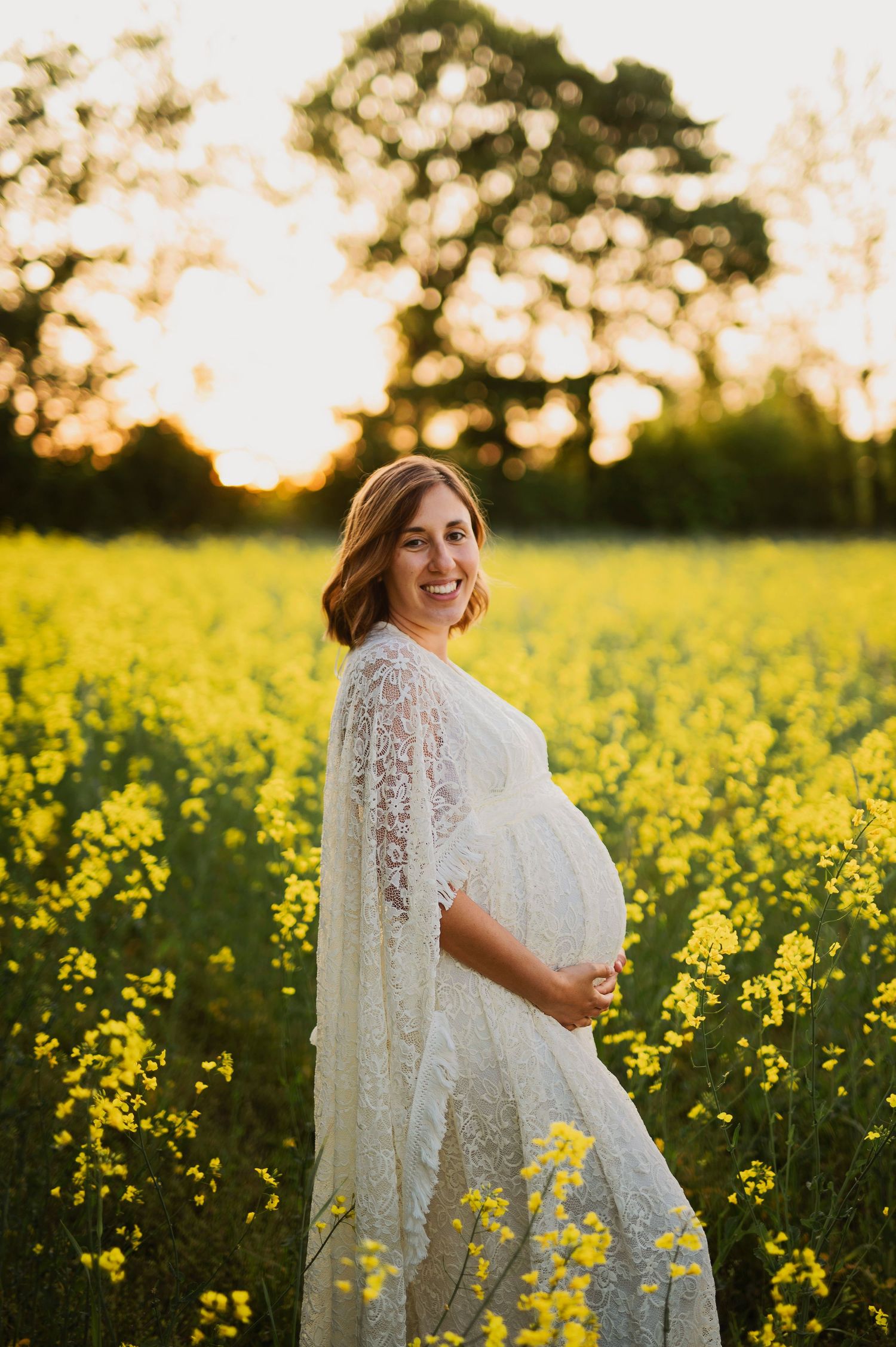 Expectant mother in white lace dress standing in field of yellow flowers at sunset.