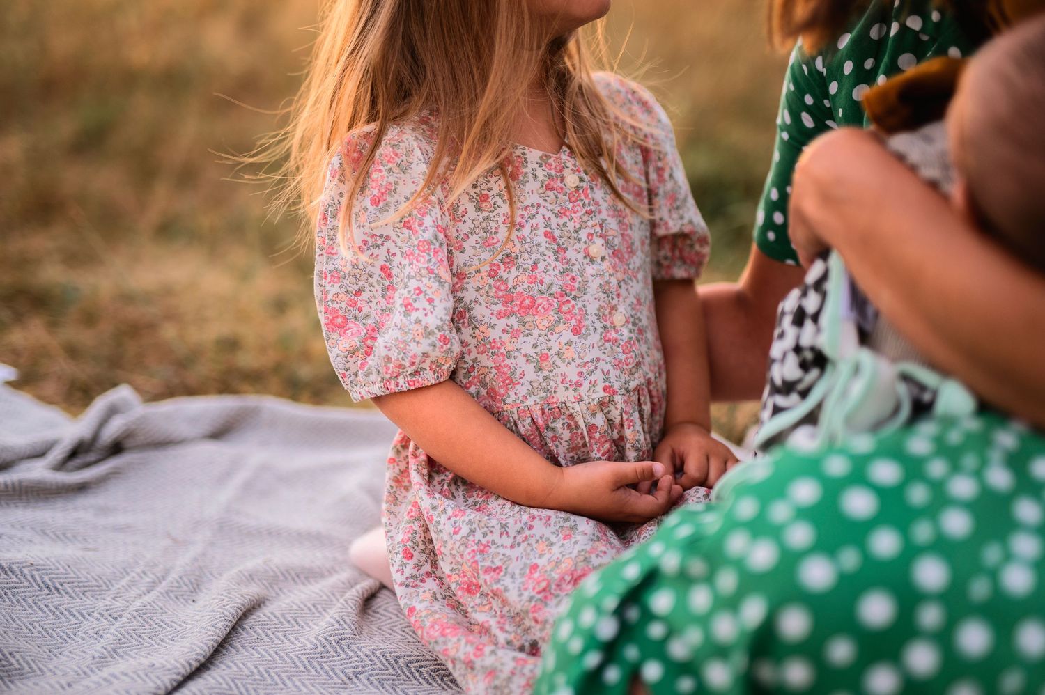 People sitting on blanket wearing floral and polka dot dresses in warm sunlight.
