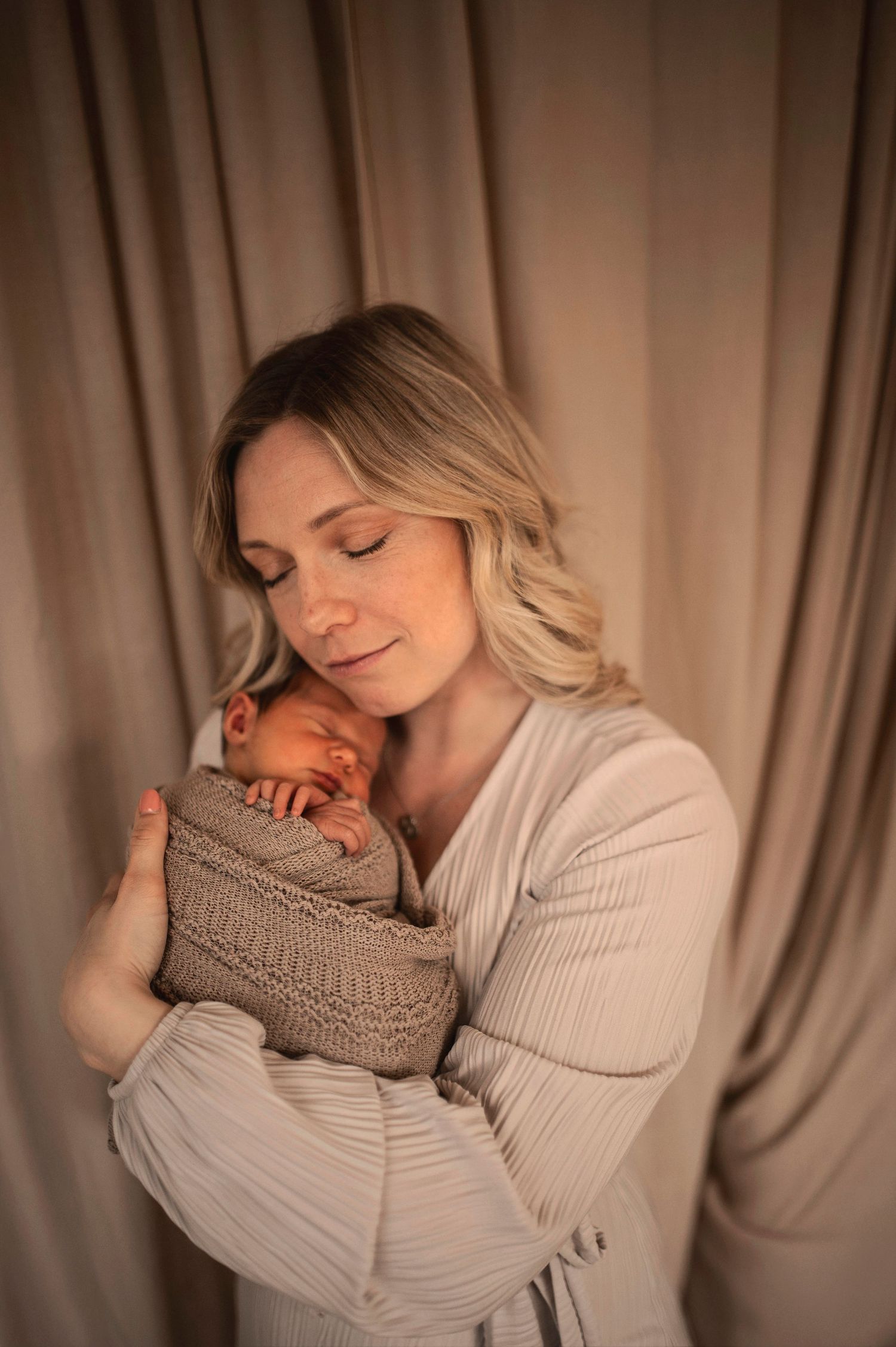A blonde mother gently cradles her swaddled newborn in warm, soft lighting.