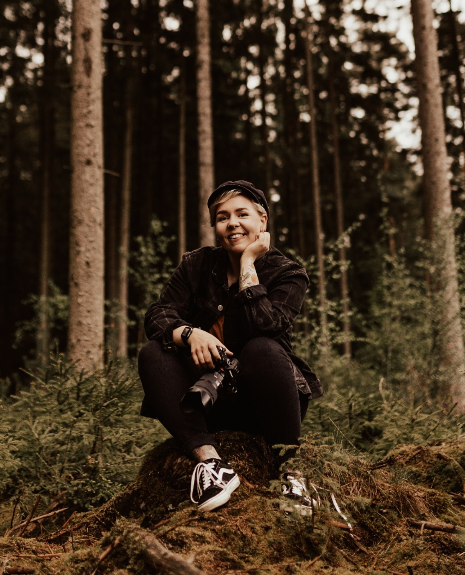 Young woman sitting on forest floor holding camera surrounded by tall pine trees.