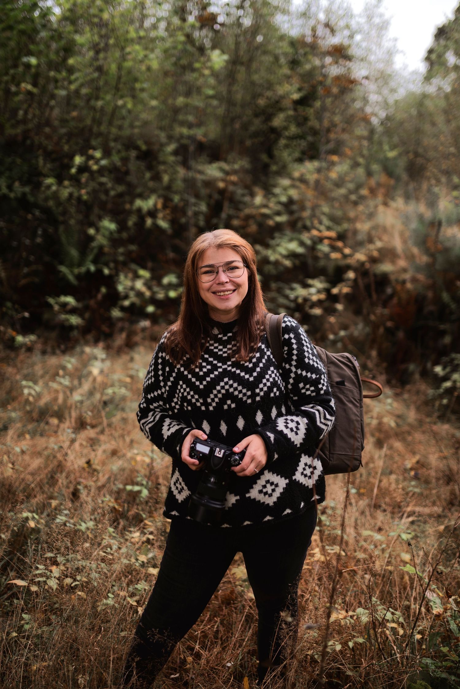 Smiling female photographer with glasses holding camera in patterned sweater standing in autumn forest field.