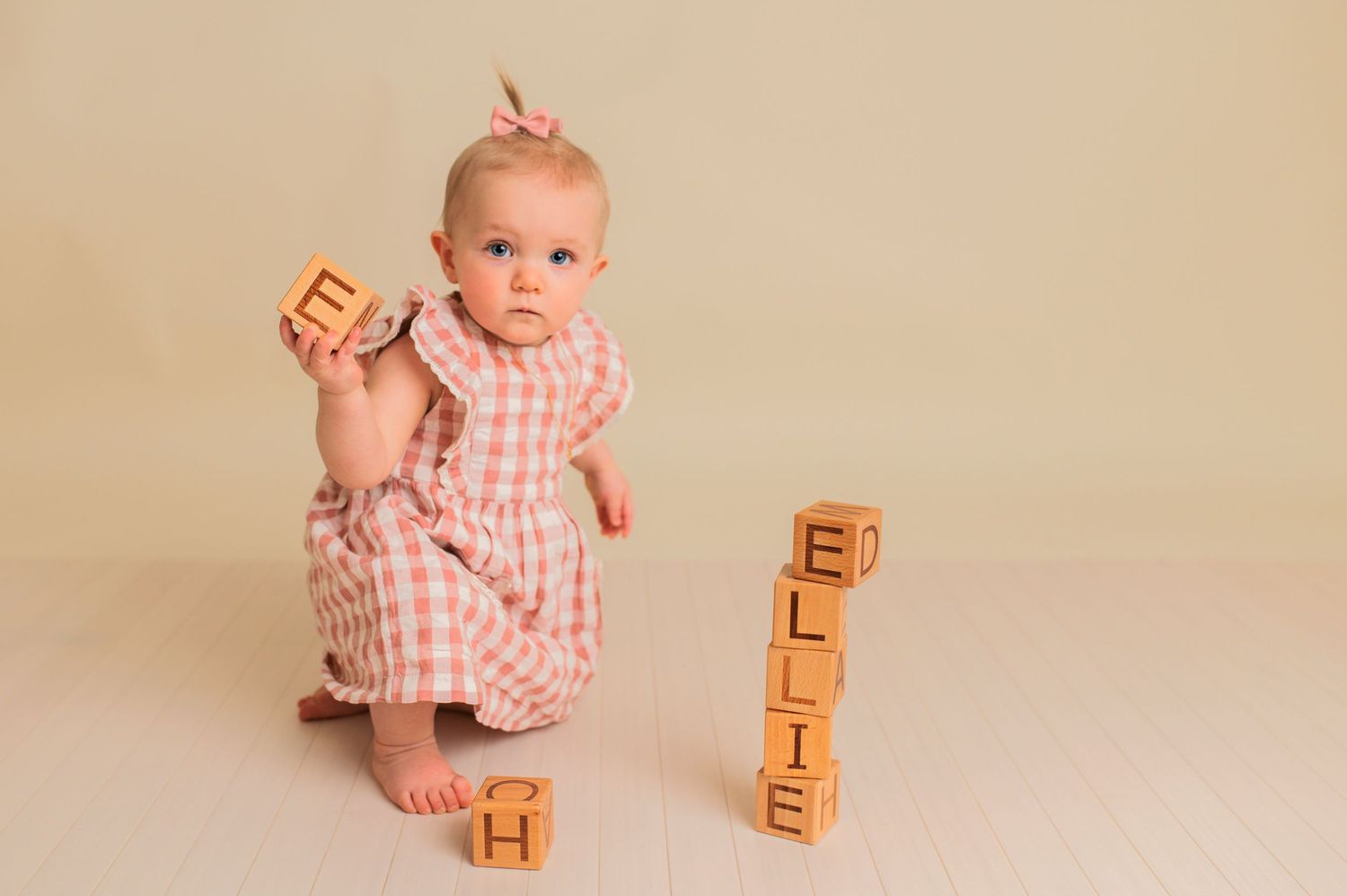 A baby in a pink gingham outfit plays with wooden alphabet blocks on a neutral background.
