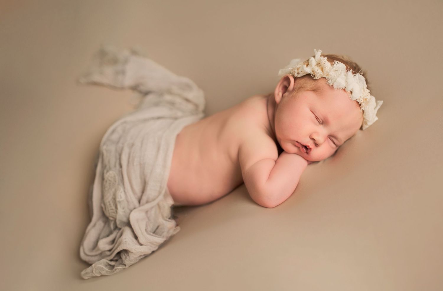 Peaceful newborn wearing floral crown lies draped in soft gray fabric against neutral background.