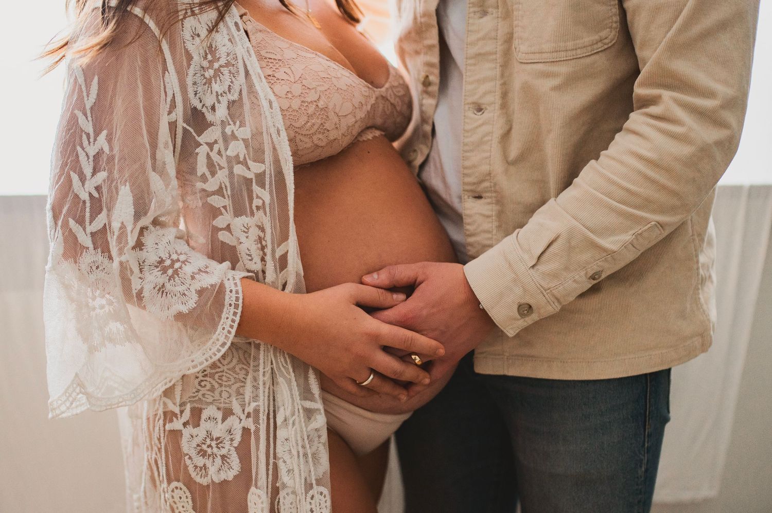 Close-up shot of expectant couple's hands resting on bare pregnant belly with lace robe details.