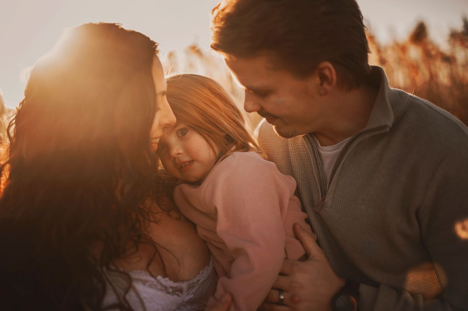 Family of three embracing during golden hour with sunlit reeds in background.