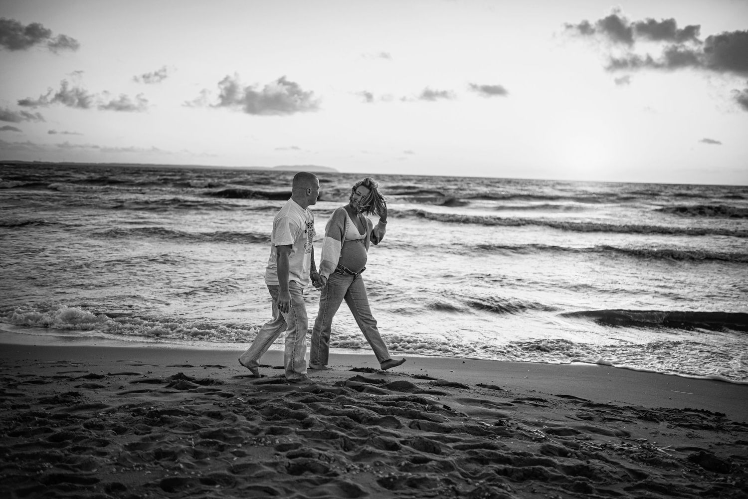 A black and white photo of two people walking along a beach shoreline at sunset with waves rolling in.