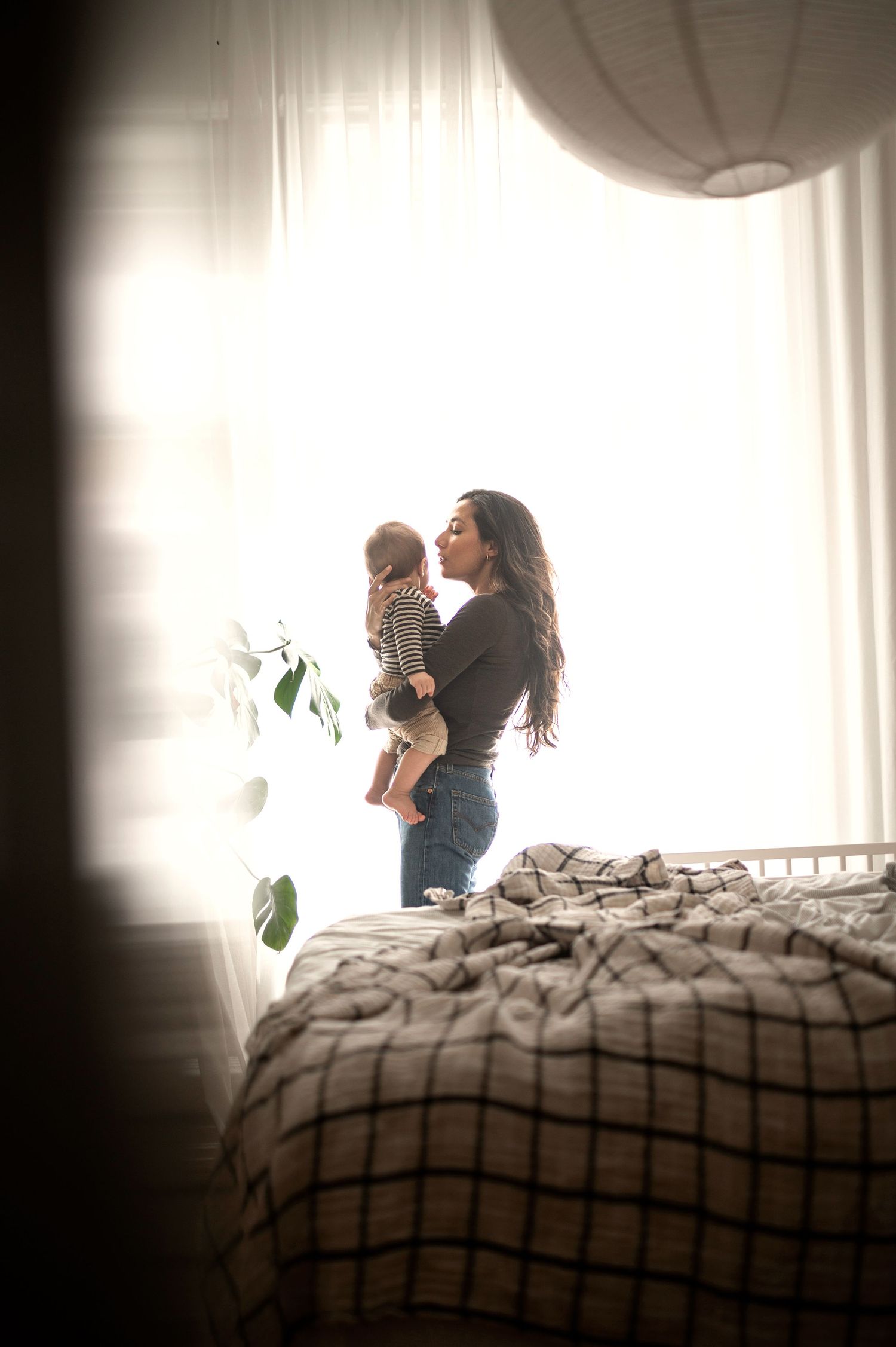 Young mother holding her baby near a bright sunlit window in a cozy bedroom.