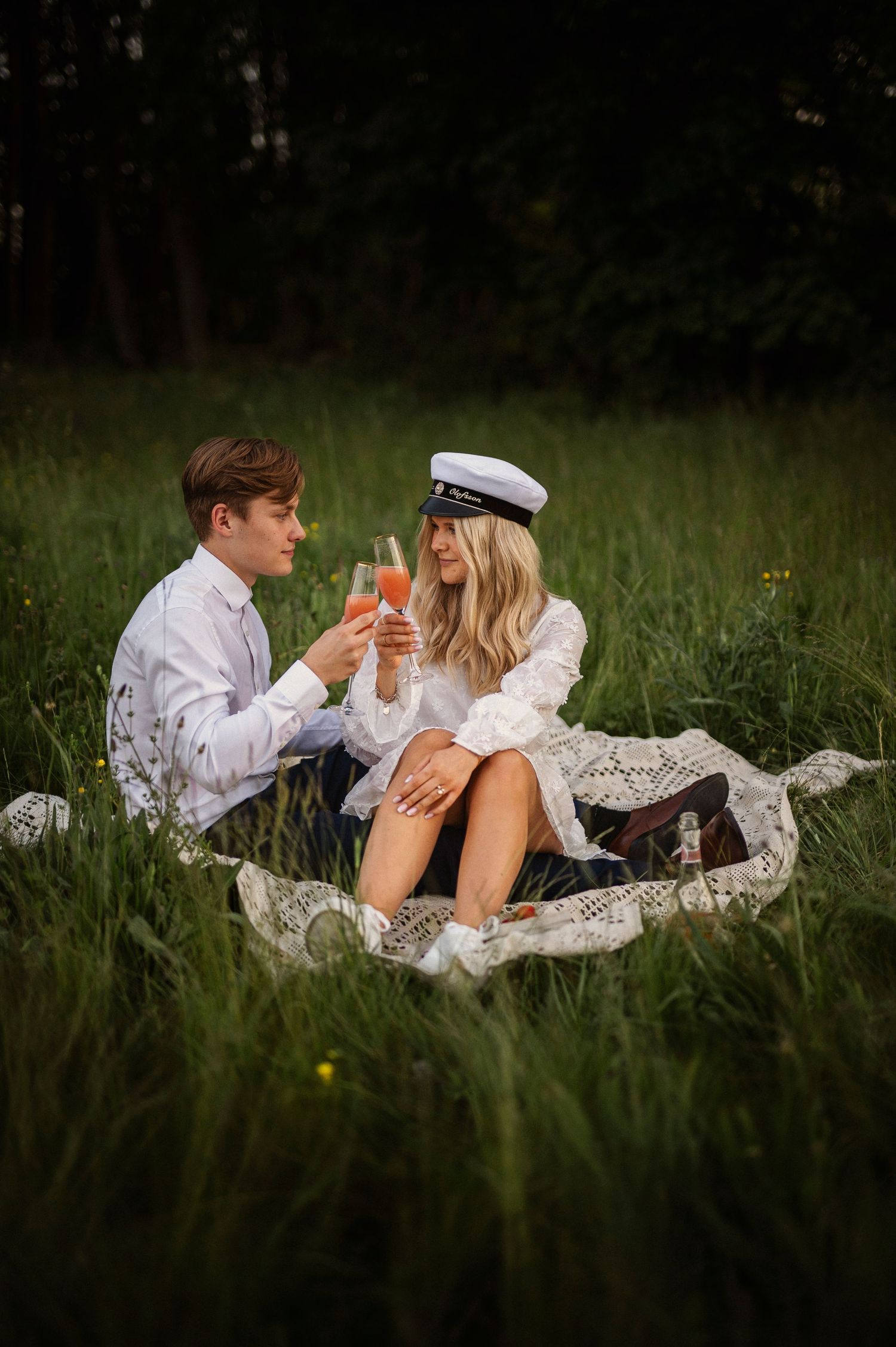 A couple toasts with pink drinks during a romantic outdoor picnic in a lush green meadow at dusk.
