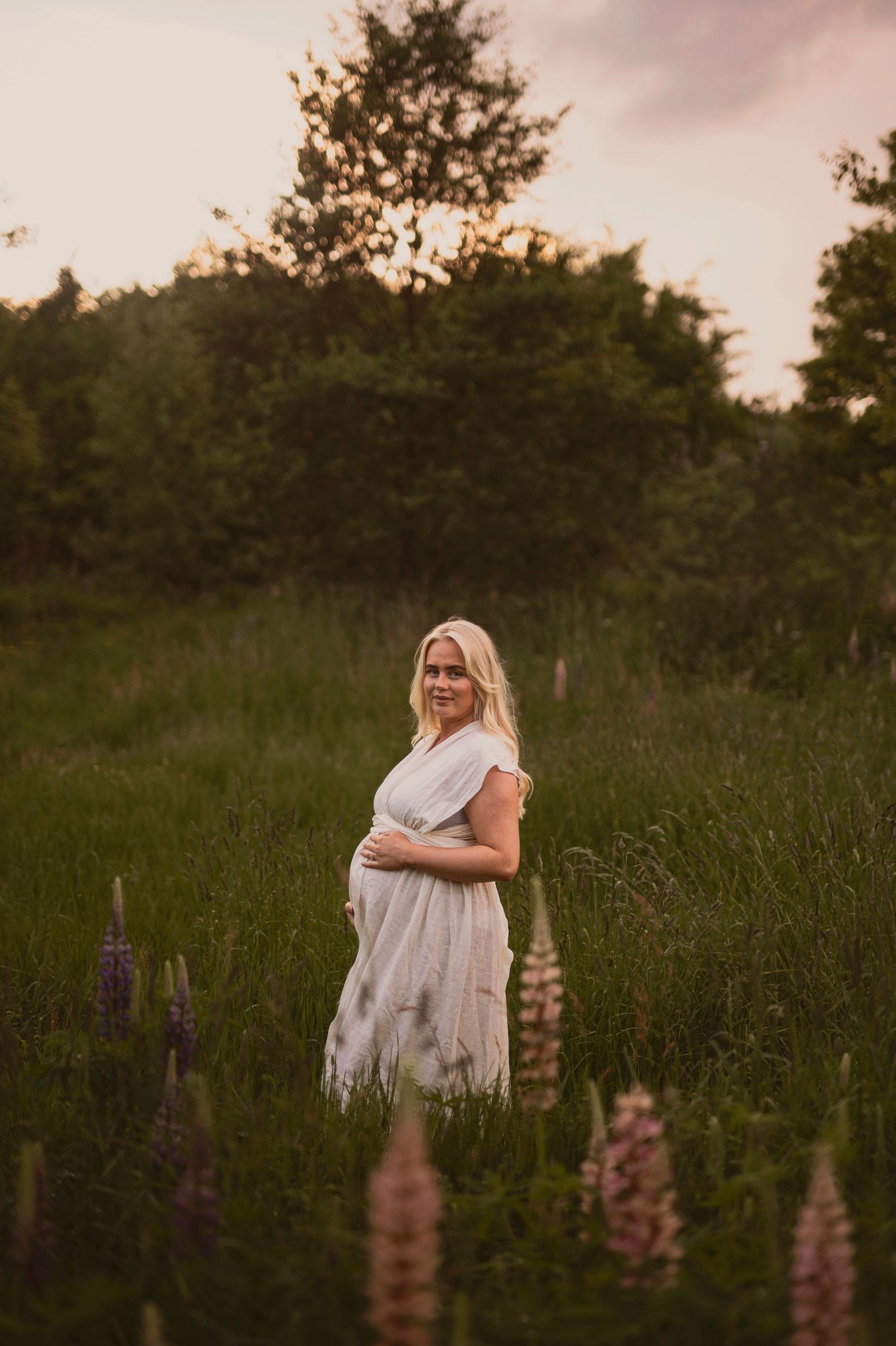A pregnant woman in a white dress stands in a field of lupine flowers at sunset.