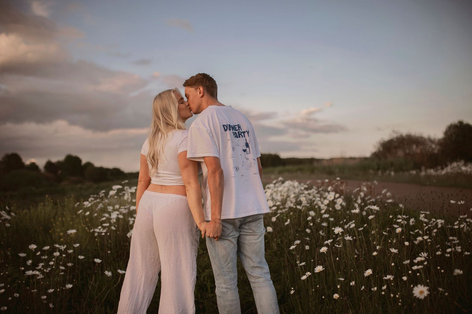 Tender maternity photography session in golden light with couple walking through field of daisies.