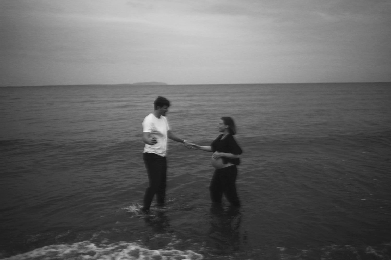 A couple stands together in shallow ocean water in black and white.
