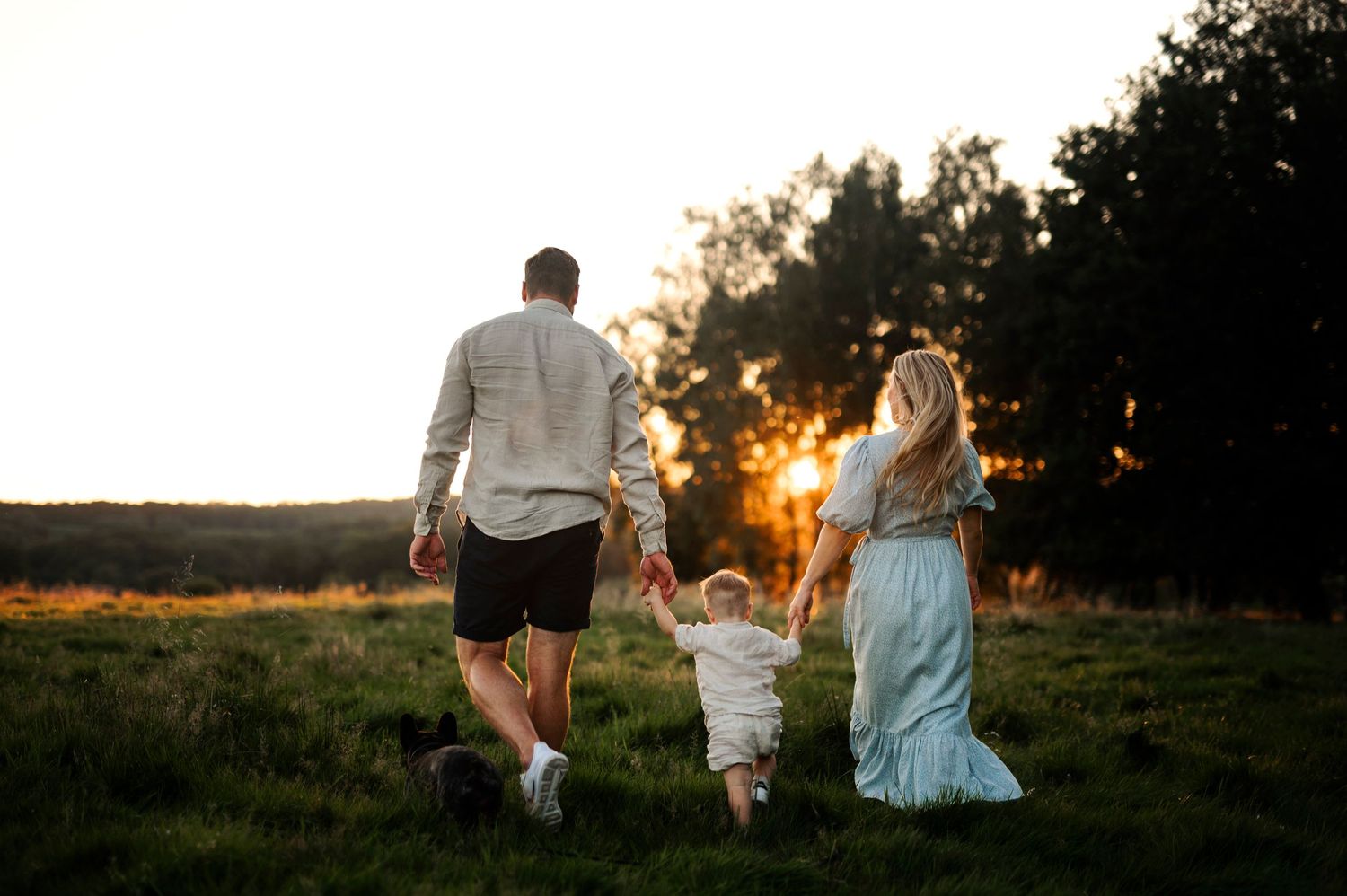 A family walks together through a grassy meadow towards a glowing sunset.