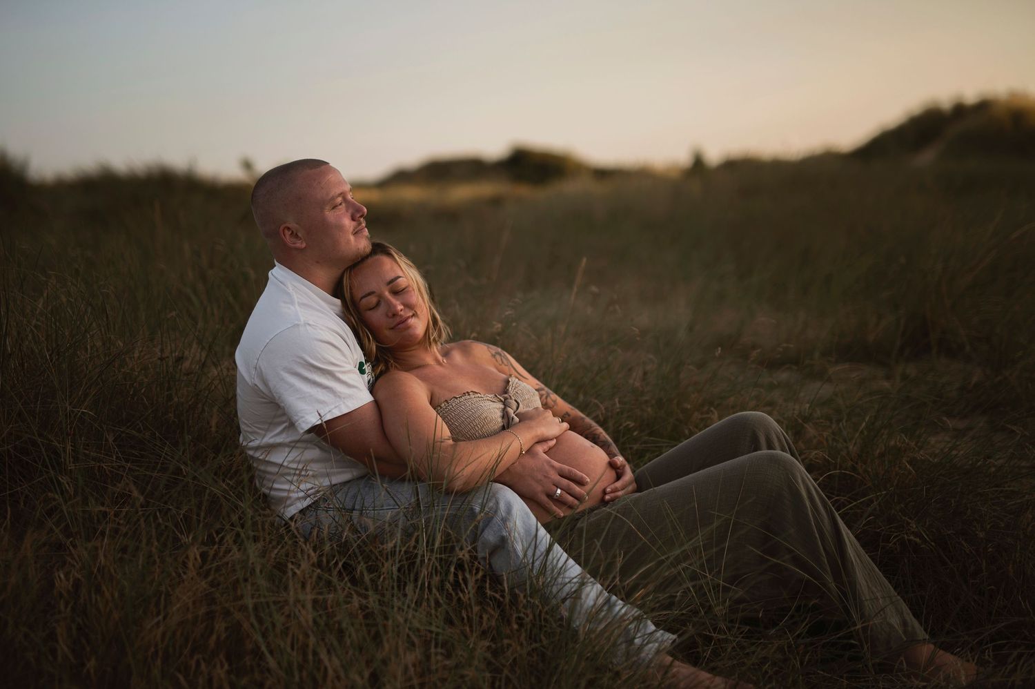 A couple embraces while sitting in tall grass during golden hour at sunset with a dreamy atmospheric glow.