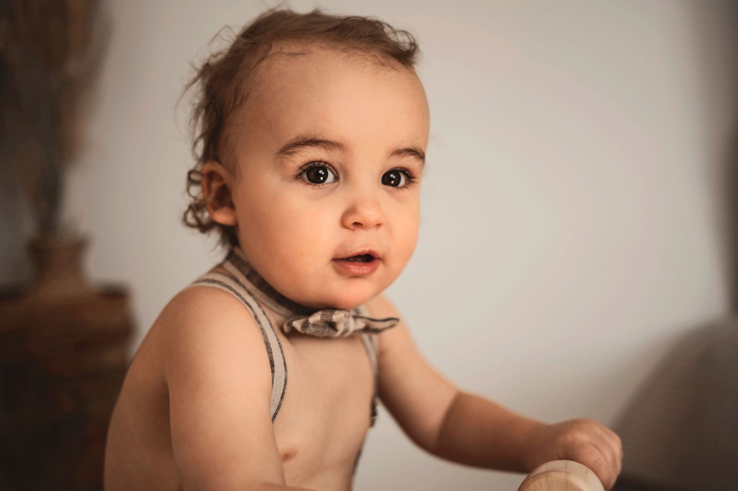 Small child in a sleeveless shirt sits with an inquisitive expression in soft portrait lighting.