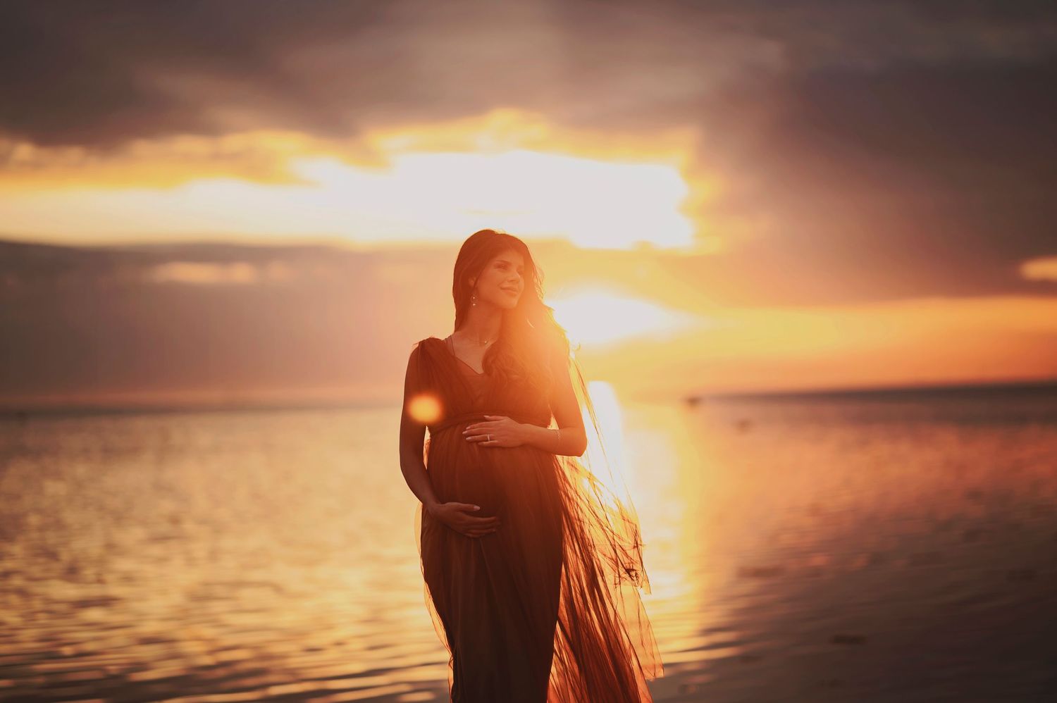 Dramatic sunset silhouette over water creates a romantic maternity portrait.
