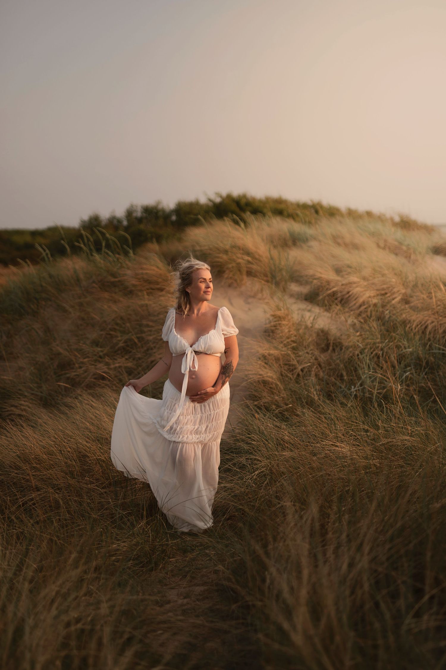 Figure in white dress stands among golden beach grass at dusk.