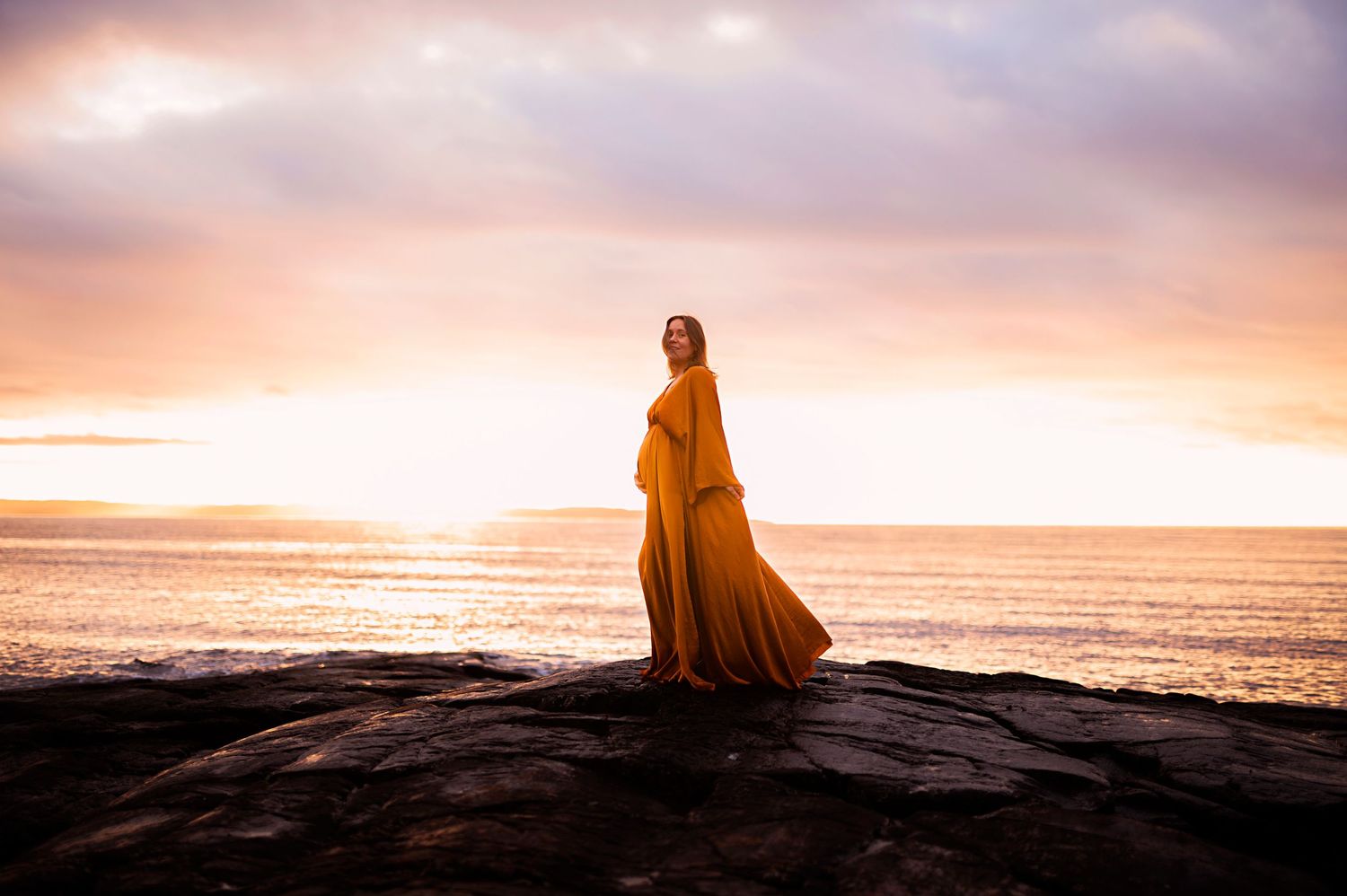 A dramatic silhouette of a figure in a flowing dress stands on rocky coastal cliffs during a golden sunset.
