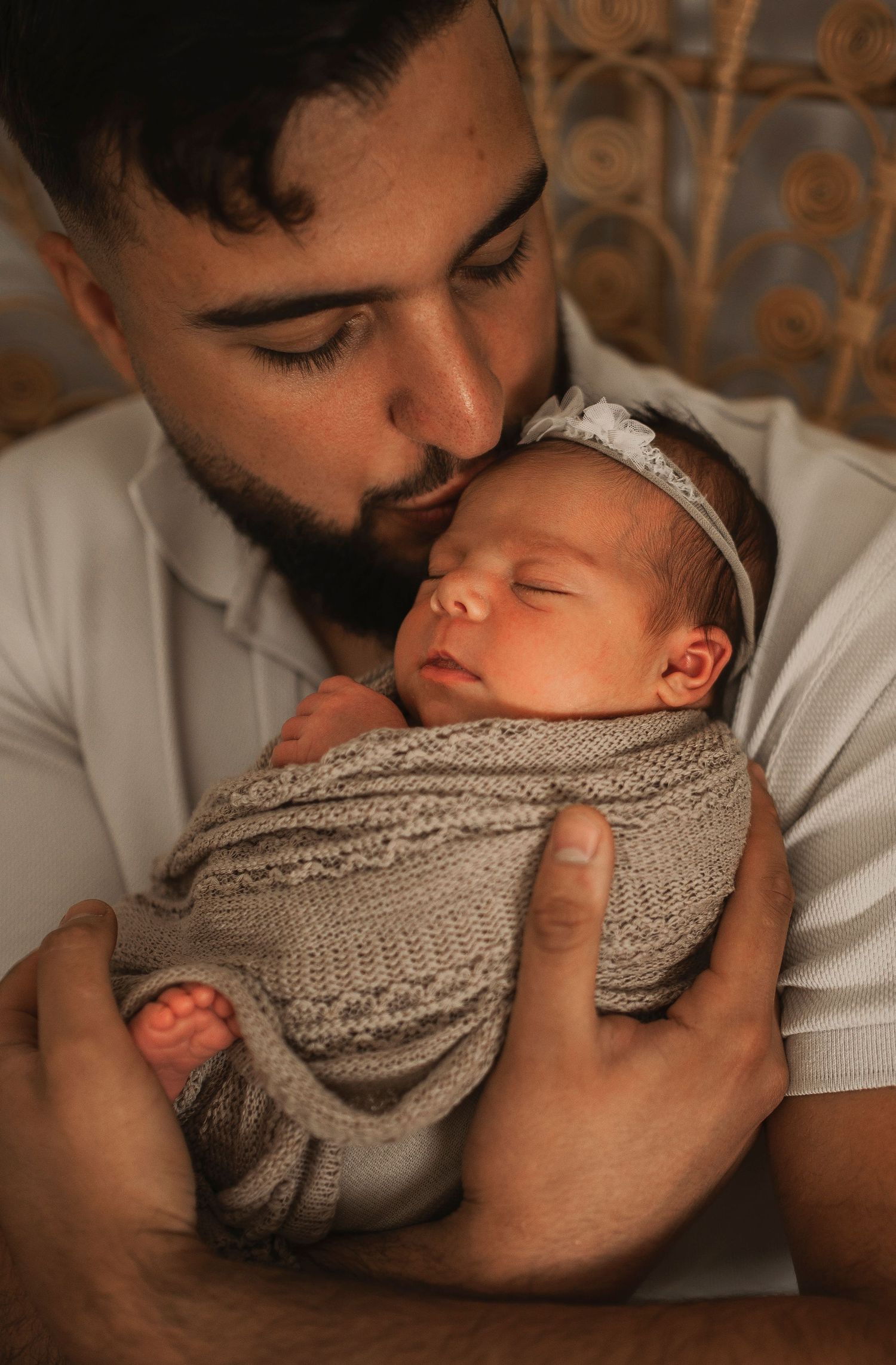 Bearded father gently kissing sleeping newborn baby wrapped in knit gray swaddle.