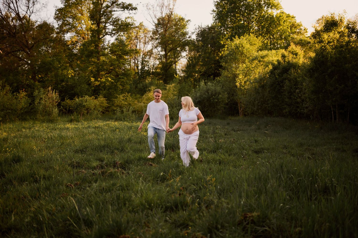 A couple in white outfits runs together through a scenic meadow surrounded by trees at dusk.