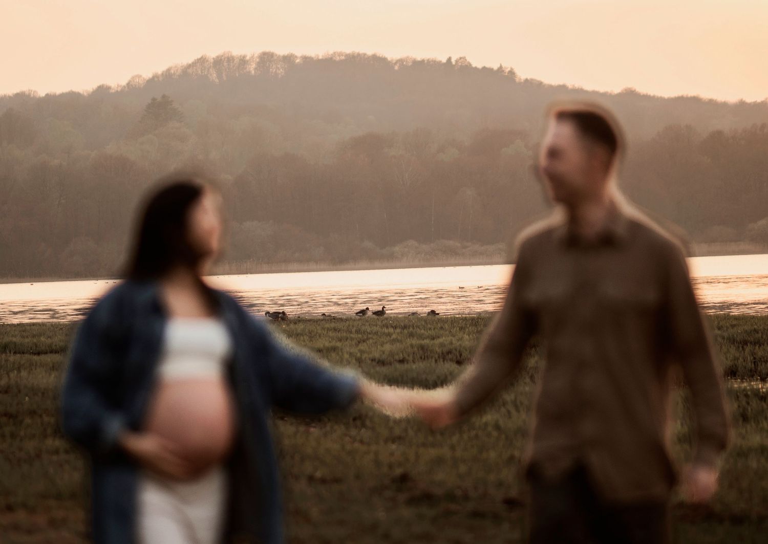 A couple holds hands while walking along a scenic lakeside at sunset with hills in the background.