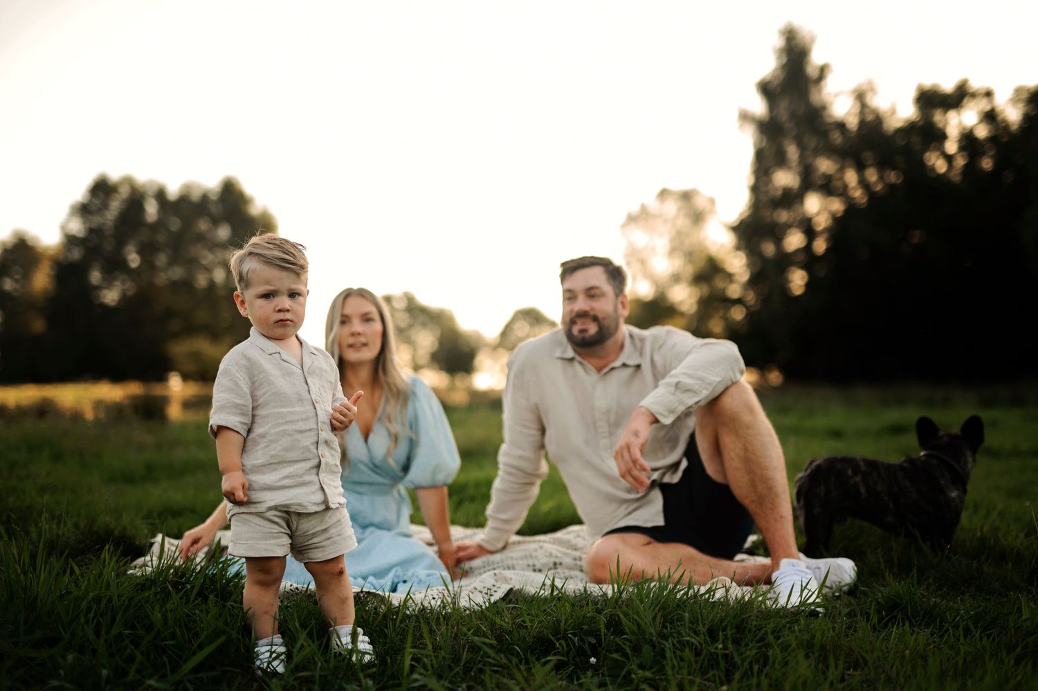 Casual family picnic scene on white blanket with black dog in grassy field at sunset.