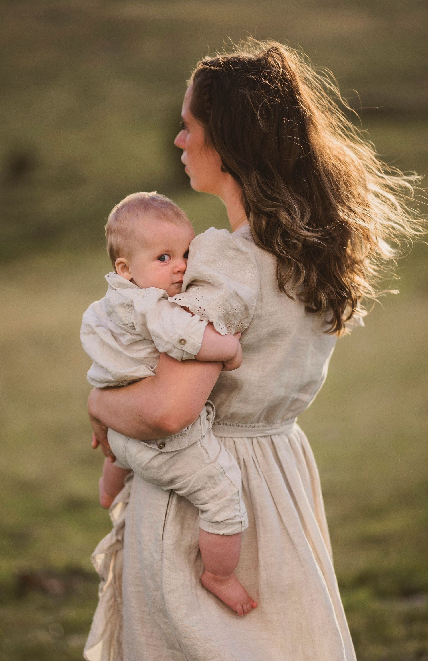 Mother in flowing dress holds baby against golden hillside backdrop as wind blows through her hair.