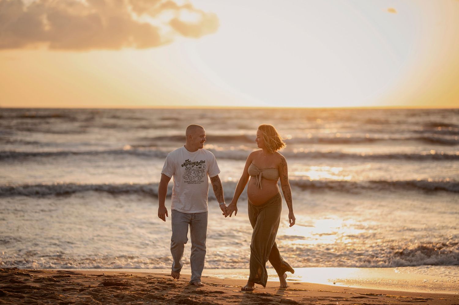 Couple walking hand in hand along beach at sunset with golden light reflecting off ocean waves.