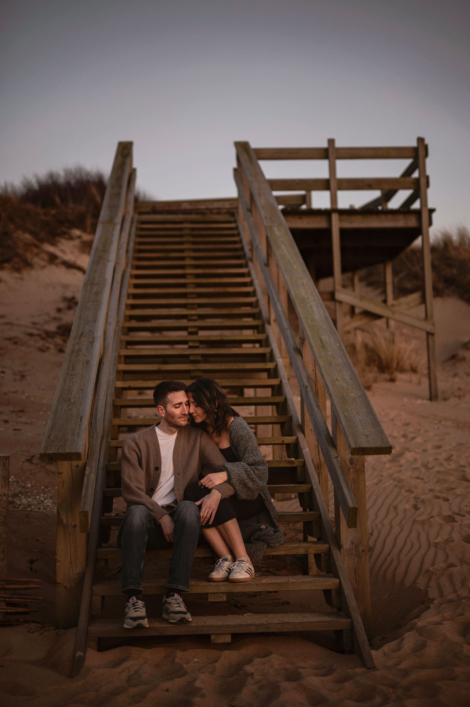 A couple sits together on wooden beach stairs at dusk, creating an intimate moment in the sand and shadows.