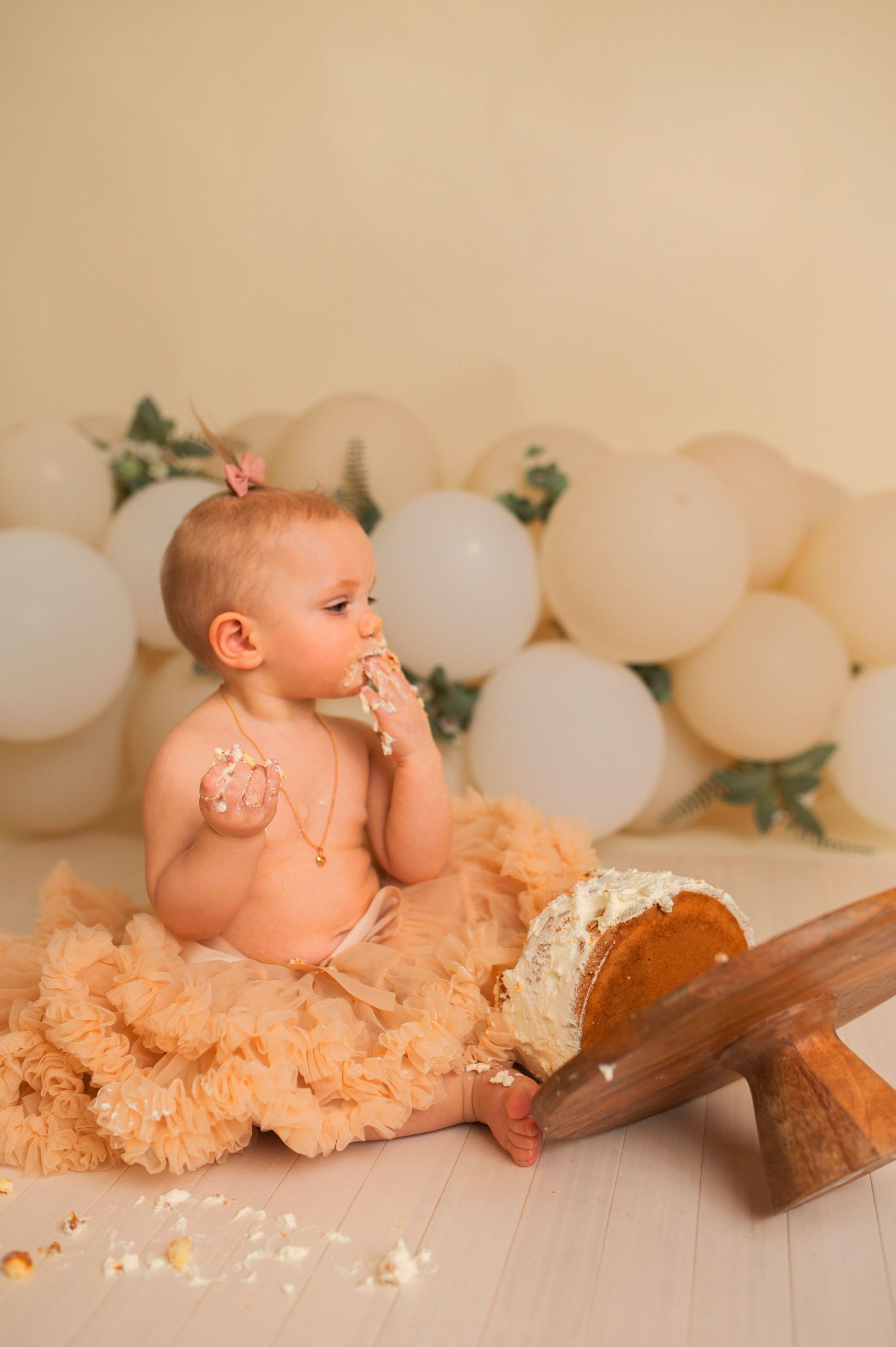 Baby in peach tutu tasting cake during first birthday photoshoot with balloon decor.