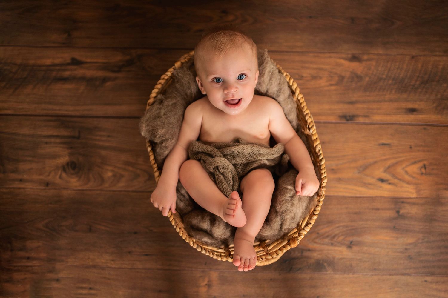 Smiling baby sitting in round wicker basket on hardwood floor.