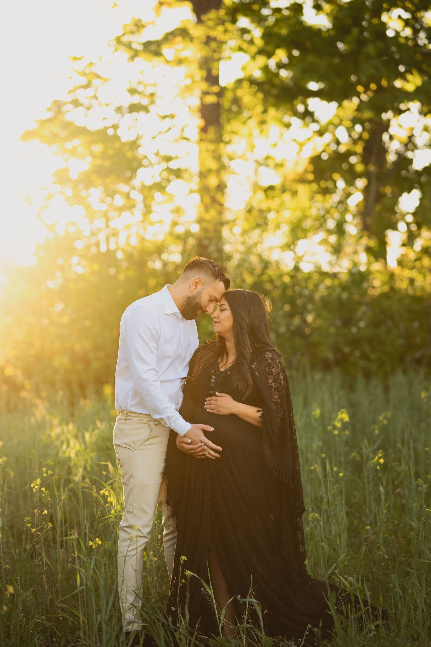 Romantic maternity photoshoot in a sunlit meadow with couple embracing in formal attire at golden hour.