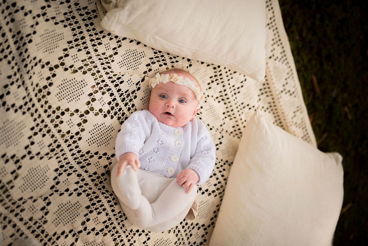 A baby wearing white clothing lies on an intricate lace blanket viewed from above.