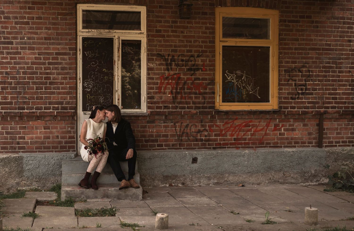 Couple sitting on steps of rustic brick building with windows.