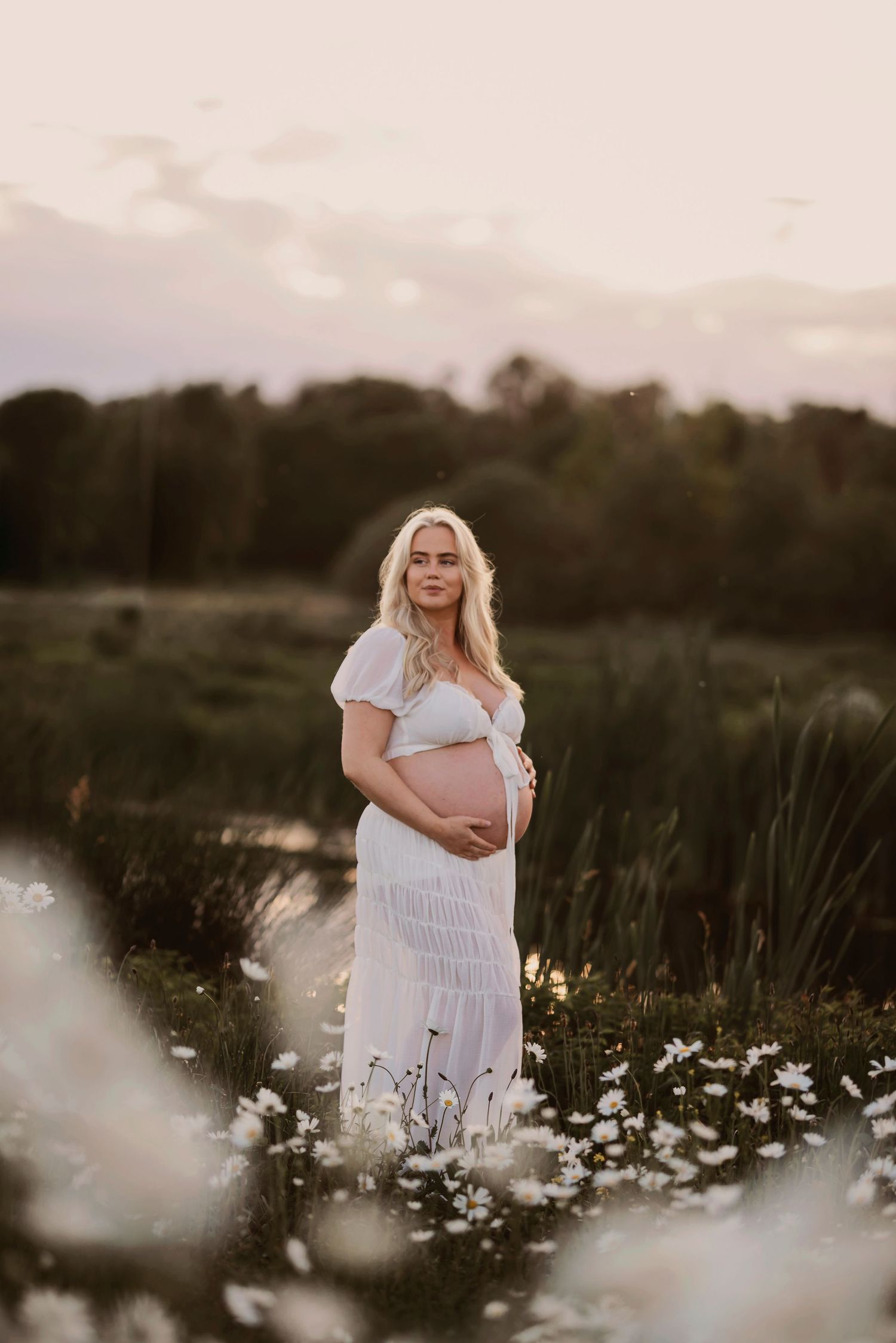 Ethereal maternity photoshoot in a dreamy wildflower meadow at sunset with white daisies and soft lighting.