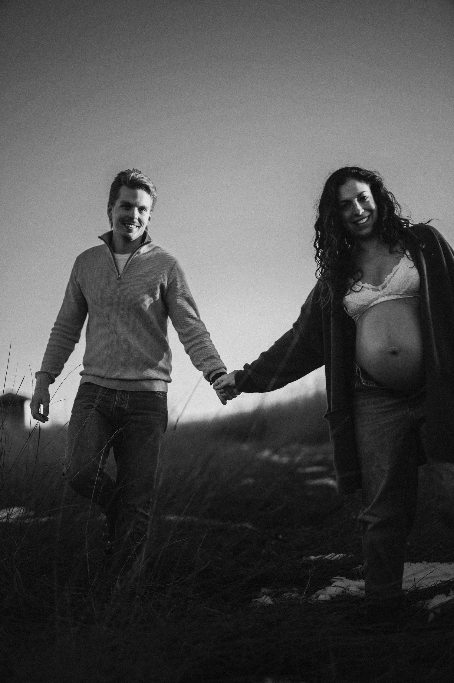 Couple holding hands while walking through a field at dusk with a pregnant belly visible.