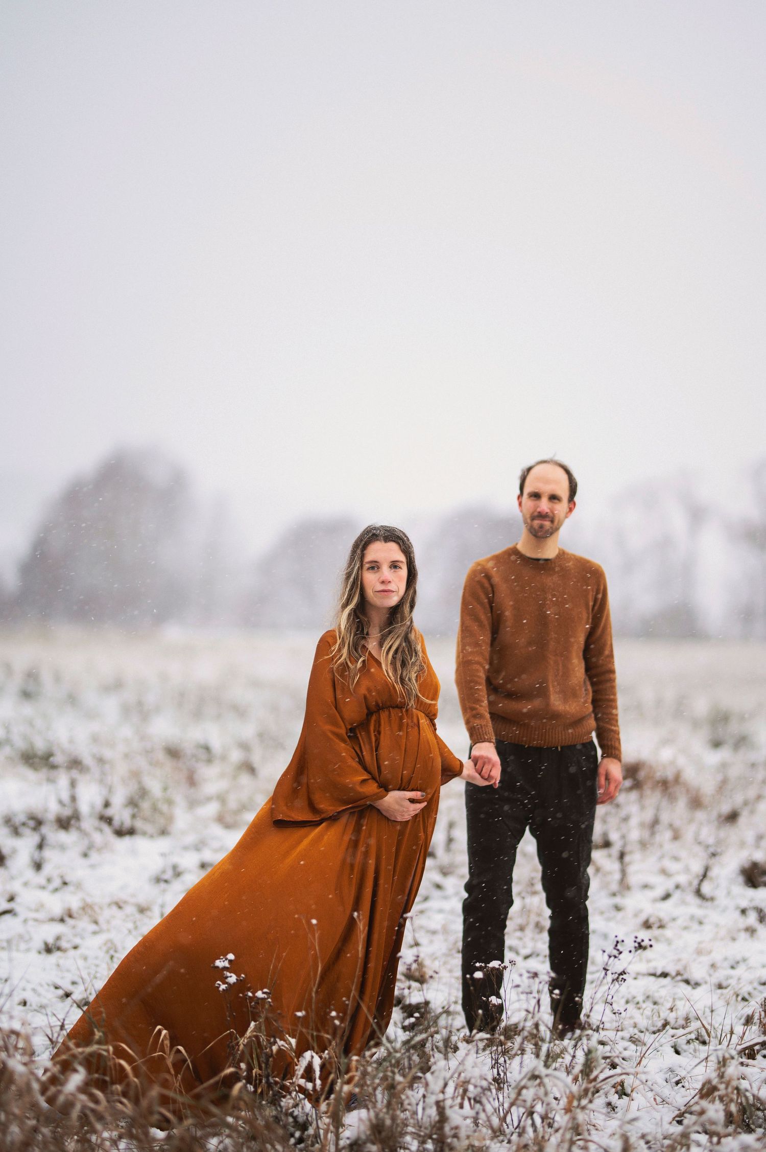 A winter maternity portrait features a couple standing in a snowy field wearing coordinating brown outfits.