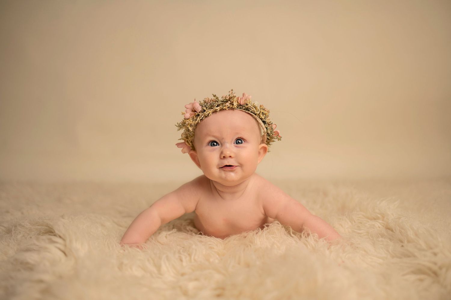 Baby wearing floral crown lying on cream colored furry rug during professional photoshoot.