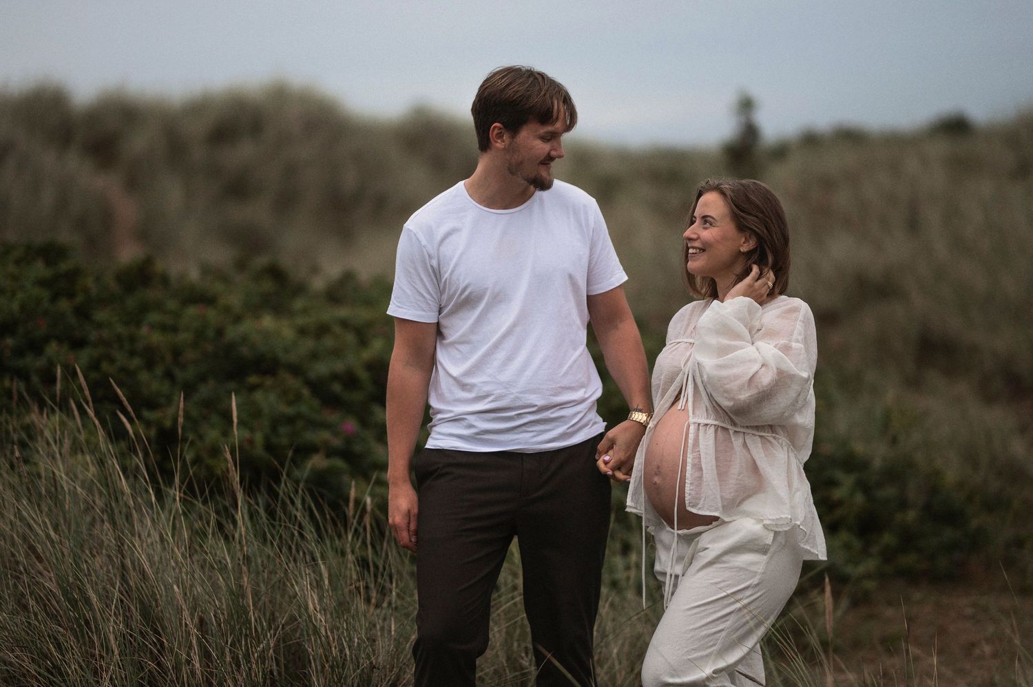 A couple stands together in tall grass on a cloudy day at the beach.