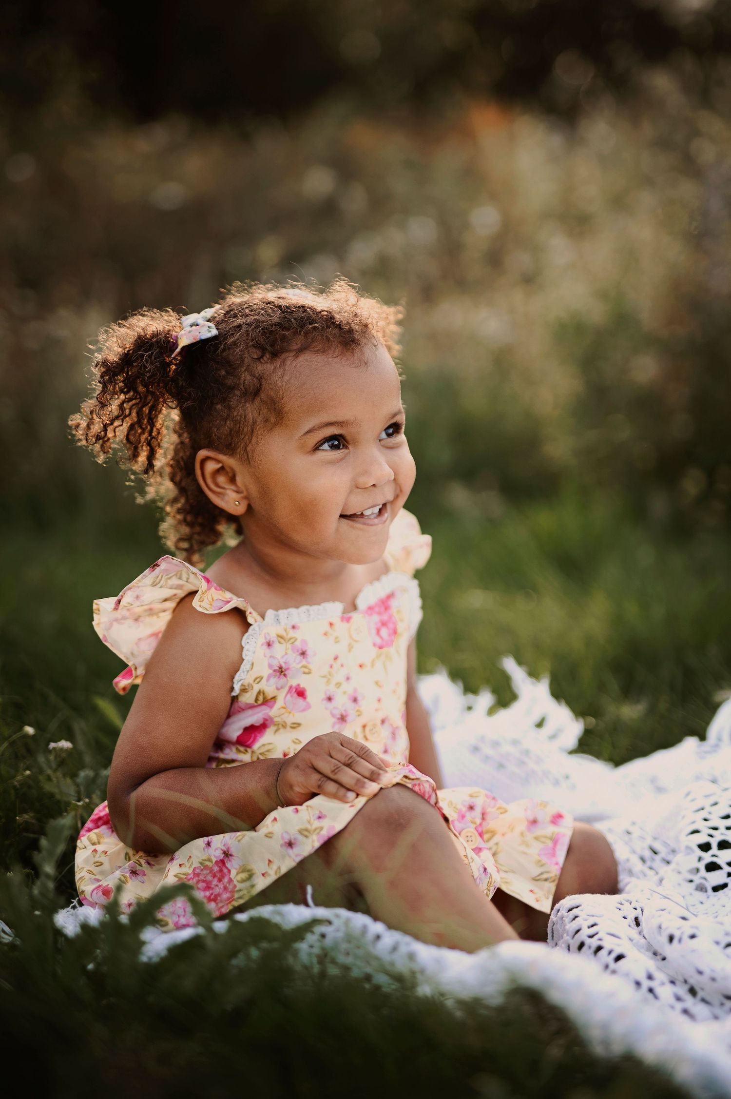A happy child sits on a white blanket wearing a floral dress in a garden setting.