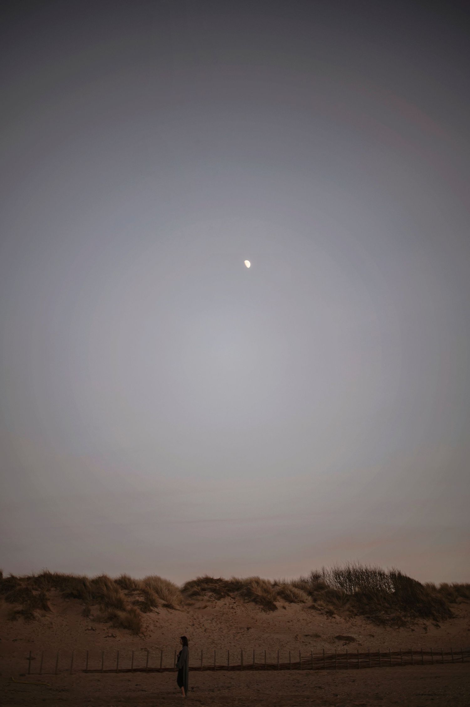 A silhouetted person stands on a beach at dusk with the moon glowing softly in the hazy grey sky above sand dunes.