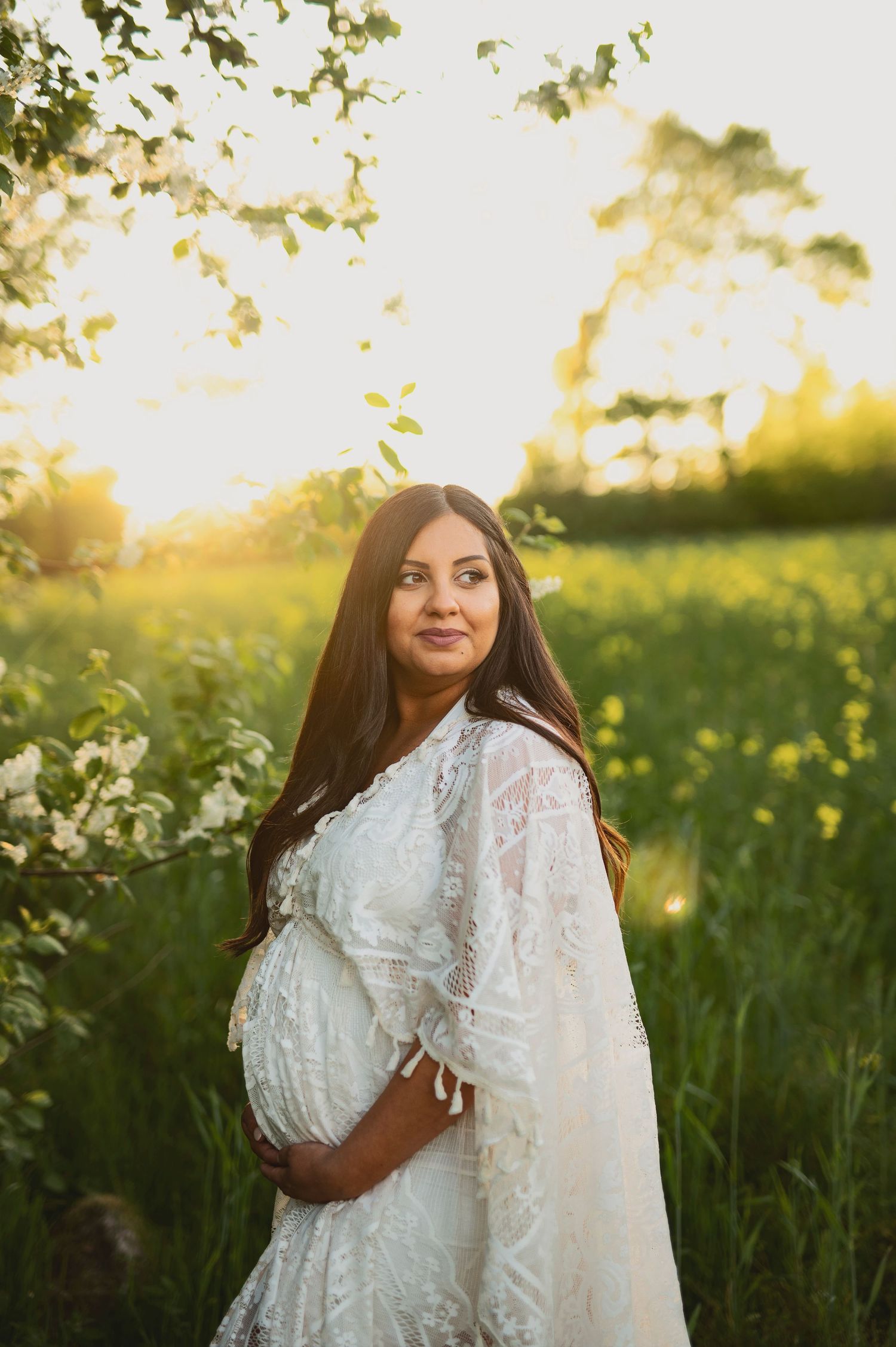 Maternity portrait of woman in flowing white dress standing amid wildflowers at sunset.