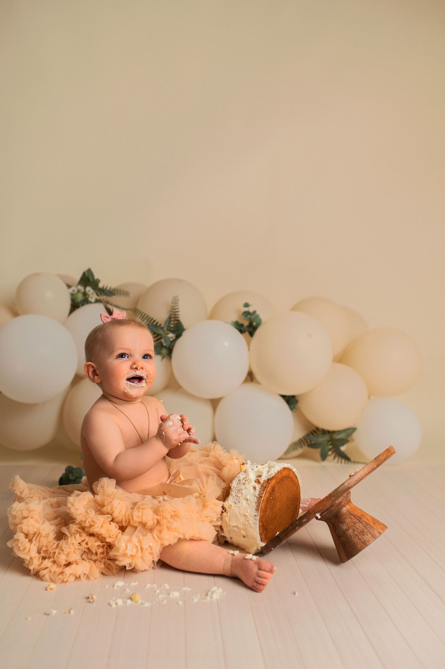 Smiling baby in peach tutu during cake smash session with white balloon and greenery backdrop.