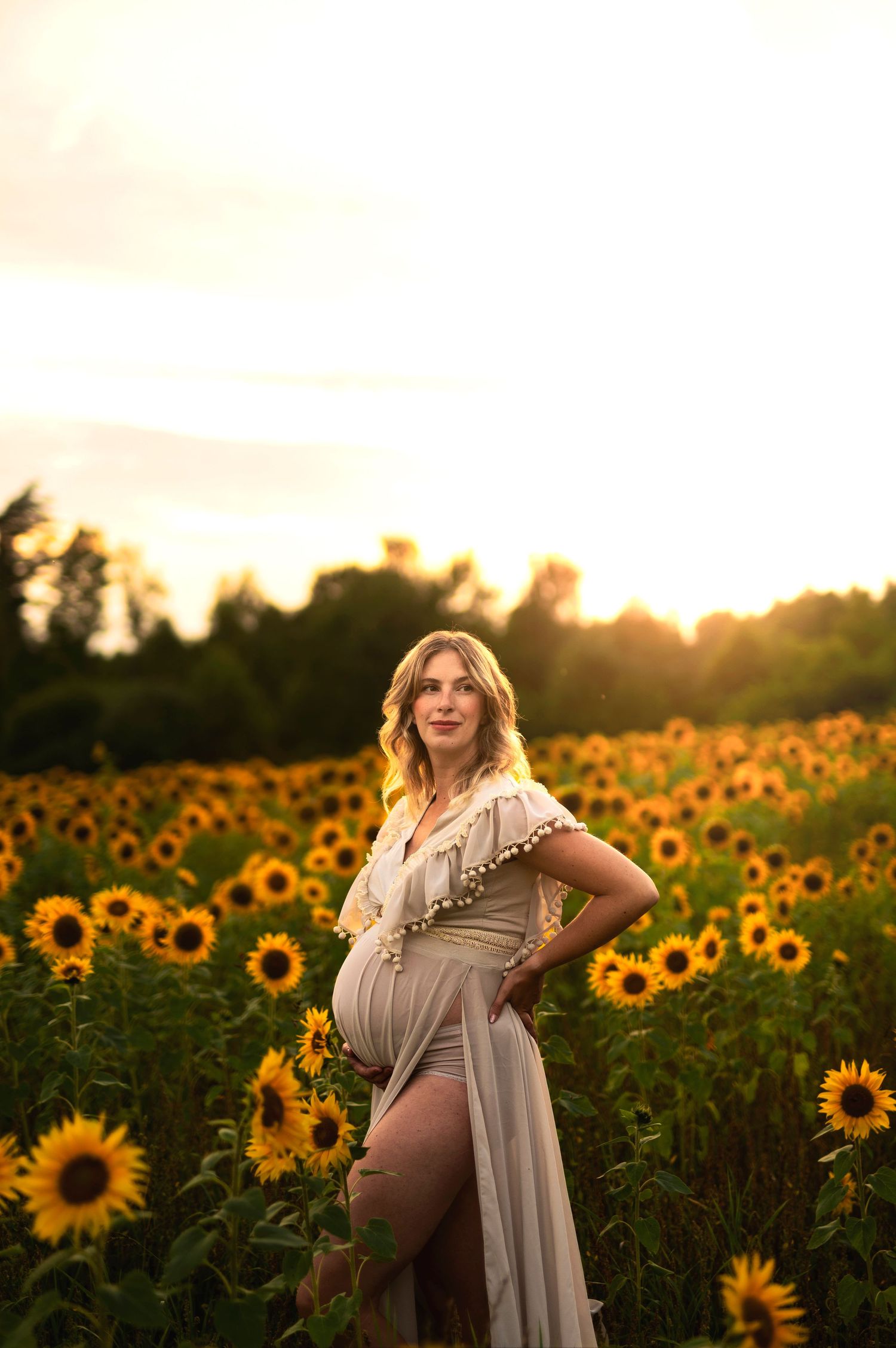 Woman in ethereal dress poses gracefully in sunflower field during sunset.